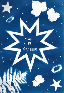 A cyanotype with a blue background and white text that says 'joy is queer'. The text is surrounded by stars and leaves.
